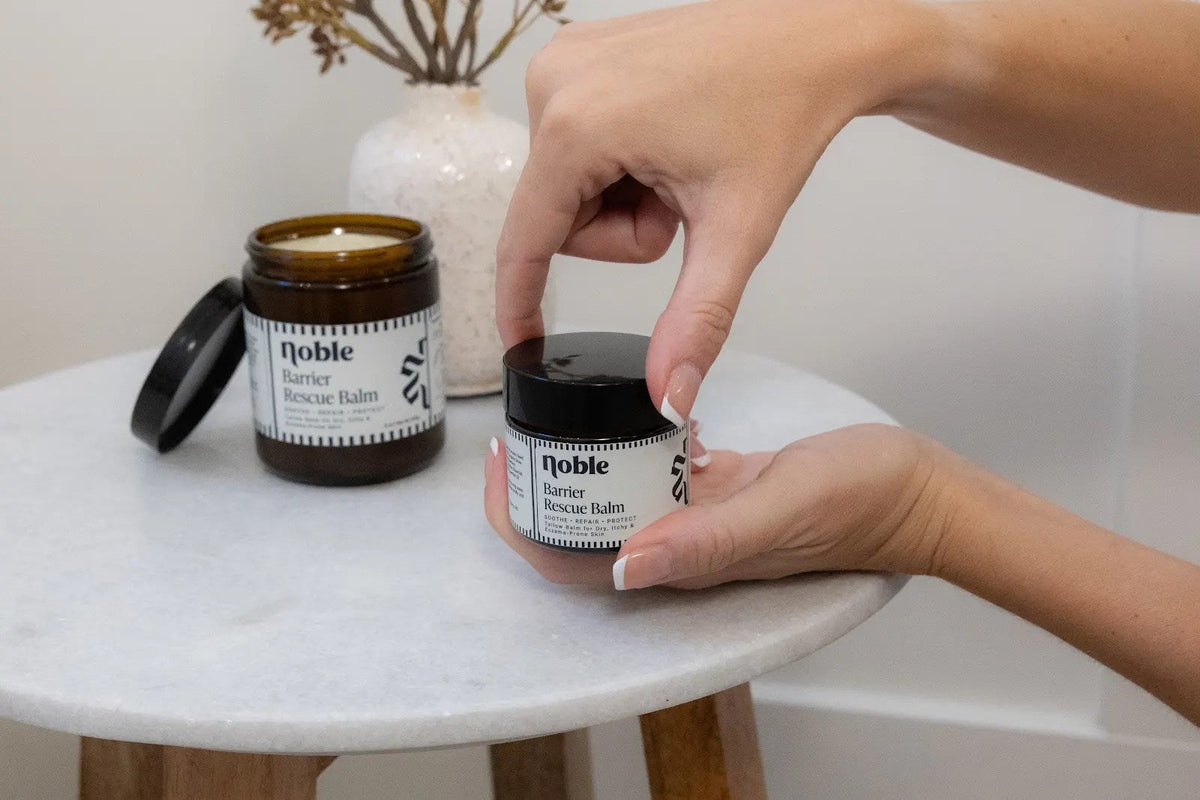 Hands holding Noble Barrier Rescue Balm beside a larger jar on a bathroom table.