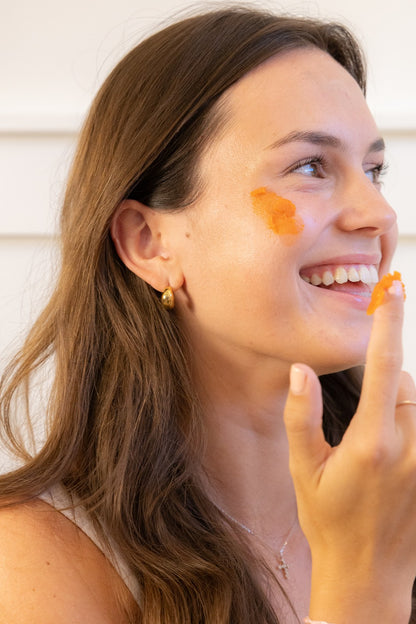 A smiling woman enjoying her skincare ritual, with a dab of balm on her cheek.