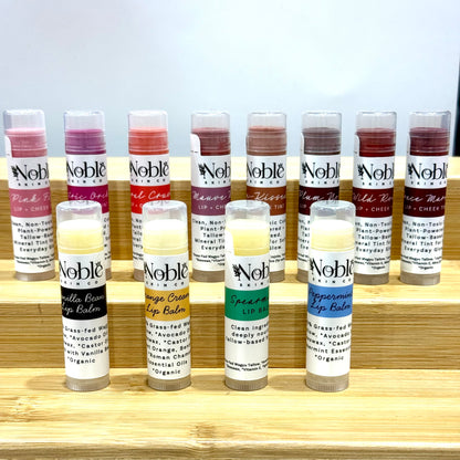 Collection of Noble Skin Co. grass-fed tallow lip balms, including tinted and untinted varieties, arranged on a wooden display.
